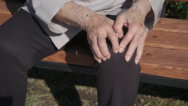 An elderly woman suffers from knee pain. An old woman in the park on a bench suffers from pain in her knees. An elderly woman rubs her sore knee.