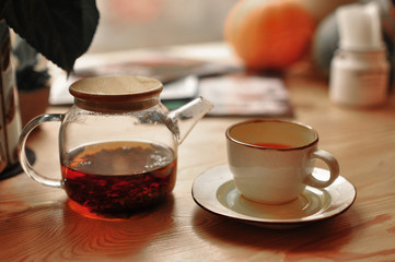 white cup of tea and saucer with a transparent teapot with a lid on a wooden table by the window