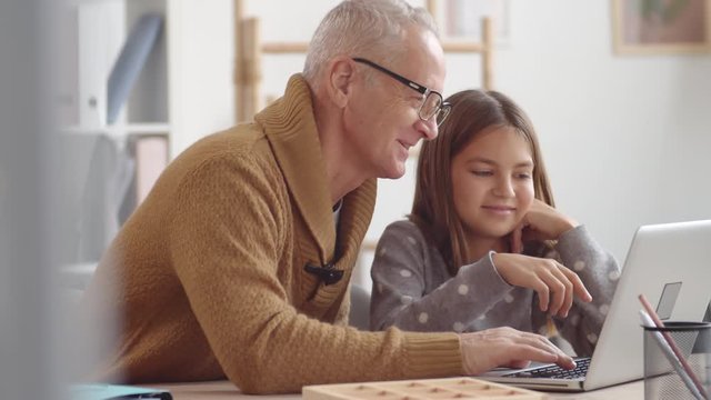 Medium shot of Caucasian beautiful girl sitting at desk and helping her grandfather wearing eyeglasses mastering PC