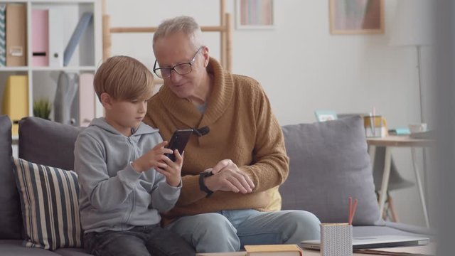 Medium shot of Caucasian boy sitting on sofa together with senior man and helping him mastering phone and smart watch on his wrist