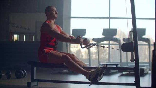 A man pulls weight to his stomach sitting in the simulator performing an exercise for the muscles of the back against the background of large Windows of the gym. Powerful male athlete doing rowing 