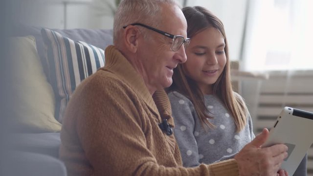 Medium shot of retired grey-haired man wearing eyeglasses and his pretty granddaughter sitting together on sofa, using tablet and having fun