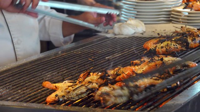 4K Close up shot of chef hand use stainless steel tongs cooking charcoal grill smoked big shrimp or prawn meat barbecue on iron sieve over the stove for self serve station in the restaurant.