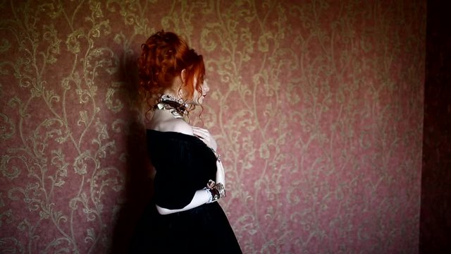 Gothic woman is a vampire with pale skin and red hair in a black dress and a necklace on her neck. Girl witch with vampire claws and red lips. Gothic look. Outfit for halloween. Model dance in room