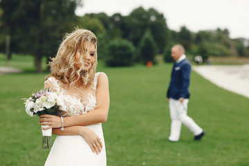 Fototapeta premium Beautiful bride in a long white dress. Handsome fiance in a blue suit. Couple in a summer park