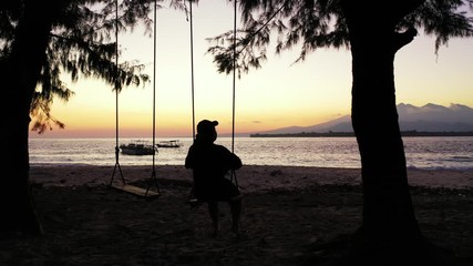 silhouette of the sad woman swinging on the swinging chair on the tropical beach on sunset.