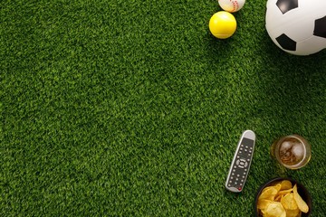 Naklejka premium Soccer ball on a green field and ottoman for a fan with snacks and a TV remote control. flat lay.Copy space.