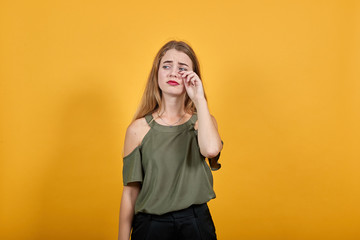 Boring young caucasian woman posing isolated on orange background in studio wearing casual shirt, keeping hand on cheek, wipes away tears