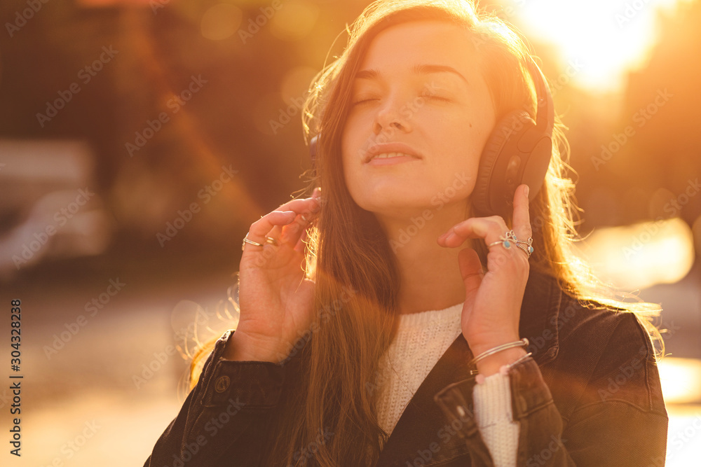 Wall mural Stylish fashionable happy hipster woman teen enjoys music in wireless headphones while walking around the city at sunset