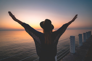 Fototapeta premium One lonely carefree enjoy traveler woman in hat with open outstretched arms standing on sea pier at sunset. Happy freedom concept and enjoying life. Free people
