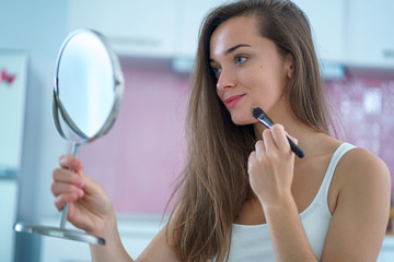 Beautiful attractive happy smiling brunette woman doing home makeup using a small round mirror and...