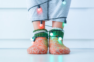 Female legs in warm knitted soft cozy christmas socks and garland with illuminated lights in...
