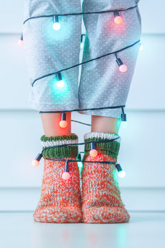 Female legs in warm knitted soft cozy christmas socks and garland with illuminated lights in wintertime at home.