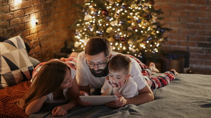 Happy father with little son and daughter in pajamas lying in bed at home, uses digital tablet computer before sleeping. Christmas tree and lights in the background