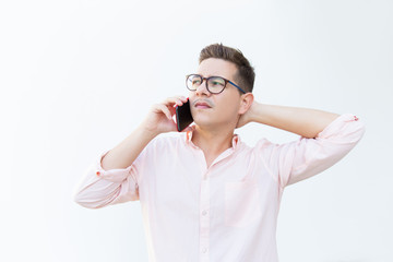 Serious pensive guy in eyeglasses touching head while talking on cell and looking away. Young man in glasses standing isolated over white background. Mobile communication concept