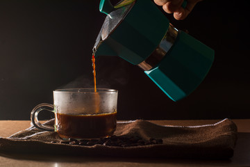 Pour hot of black coffee from green moka pot to clear coffee cup with smoke and coffee beans on...