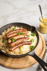 Kleine Bayrische Nürnberger Rostbratwürste mit Sauerkraut, Kartoffelpüree und Senf in rustikaler Gusseisenpfanne auf Holz Brett und Marmor Tisch