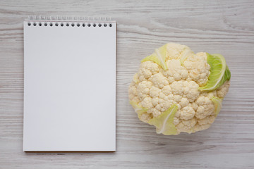 Raw Cauliflower Head, blank notepad on a white wooden surface, top view. From above, overhead, flat...