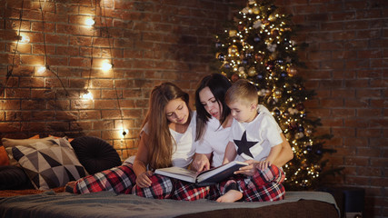 Mom with two children in pajamas sitting on the bed on Christmas night, a woman reading a book to children, in the background a garland on the wall and a Christmas tree with lights