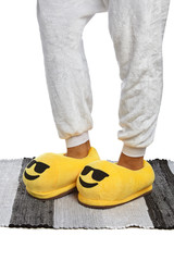 Close-up shot of female legs in yellow plush house slippers made as cool emoji in sunglasses. The girl is standing on the striped gray and white carpet.