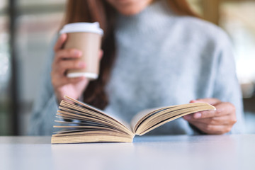 Closeup image of a woman holding and reading a book while drinking coffee on wooden table
