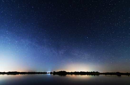 A magical starry night on the river bank with a large tree and a milky way in the sky and falling stars in the summer.