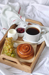Breakfast on white bed sheets in hotel room. Close up of wooden tray with coffee, bun, jam and grape