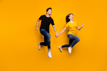 Fototapeta na wymiar Full size photo of sporty guy and lady couple jumping high holding hands pair speed marathon participants wear casual jeans t-shirts isolated yellow color background