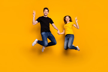 Fototapeta na wymiar Full body photo of crazy guy lady couple jumping high holding arms showing v-sign symbols saying hi wear casual jeans black t-shirts isolated yellow color background