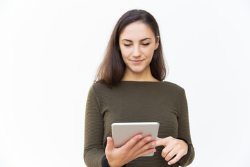 Fototapeta premium Focused positive beautiful woman using tablet, touching screen and looking at screen. Young woman in casual with digital device standing isolated over white background. Online connection concept