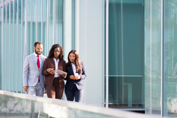 Fototapeta premium Three smiling coworkers looking at tablet during stroll. Cheerful business partners walking on street during sunny day. Business concept