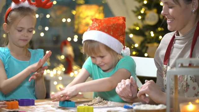 Woman and little girls baking Christmas cookies. Children with their mom making gingerbread for family dinner on Christmas eve in dining room with christmas tree and candles.