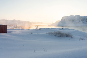 Fototapeta na wymiar snow-clad shore of the bay lit by the rays of the orange sun at dawn beyond the Arctic Circle. dense fog rises from the water on a frosty winter day
