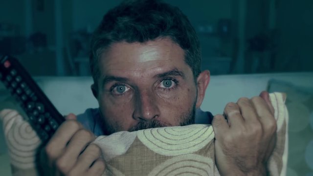 domestic lifestyle shot on excited and scared man at home living room sofa watching horror movie on television holding TV remote screaming in panic and fear at some shocking and frightening scene