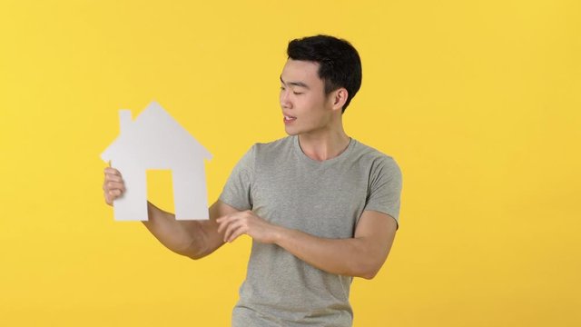 Young Asian man in gray t-shirt looking at and pointing to house cutout model while acting surprised or pleased against yellow background
