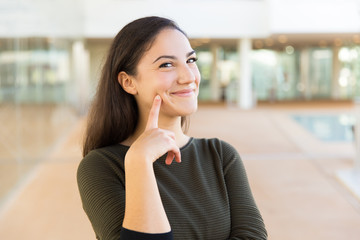 Fototapeta premium Happy cheerful beautiful woman touching face with index finger. Young woman in casual posing indoors with glass wall interior in background. Skin care or female portrait concept