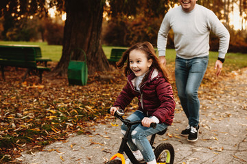 Lovely little girl going ahead with the bicycle laughing while her father is running outdoor.