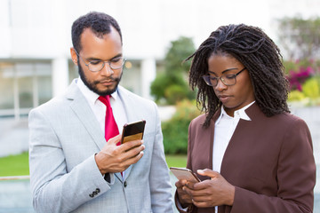 Two focused businesspeople using smartphones. Concentrated African American people holding phones while standing on street. Technology concept