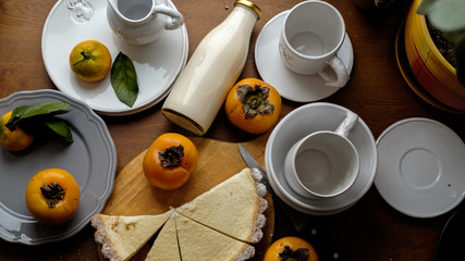 sour cream cake with persimmon and tangerines on a wooden table, next to a bottle of farm milk and a cup