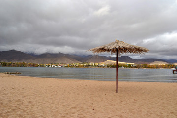 A lonely beach umbrella made of reeds on a empty beach. In the background there are gray clouds over the village and the mountains. Bad weather on vacation. Closing of the beach season, Kyrgyzstan