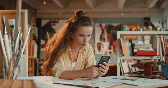 Young Caucasian pretty woman painter sitting at the table in the cozy workshop, tapping and texting on the smartphone.