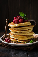 Stack of homemade little pancakes with honey, fresh raspberries and red currants on an old wooden background. Selective focus.