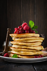 Stack of homemade little pancakes with honey, fresh raspberries and red currants on an old wooden background. Selective focus.