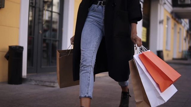  lose up of woman legs wearing stylish black coat and jeans at day time hold colored shopping bags after big shopping day. Fashion shopper shopaholic in slow motion