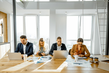 Group of a young office employees dressed casually in the suits having some office work at the large meeting table in the bright sunny room