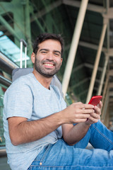 Smiling young man sitting on floor at airport. Cheerful traveler with smartphone looking at camera. Travel concept