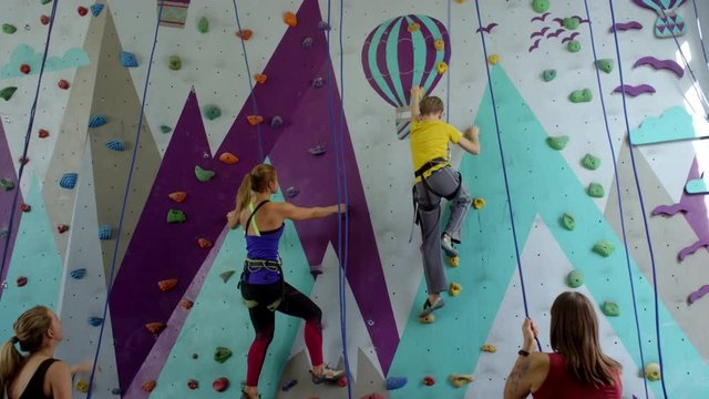 Wide tracking shot of teenage boy and young woman in safety gear, with ropes climbing up artificial wall at indoor gym, and their companions standing on ground and belaying