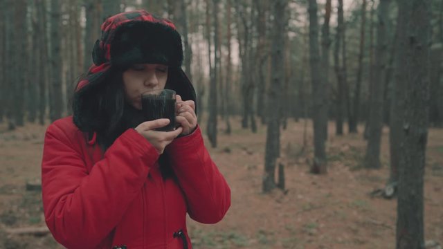 A portrait of the pretty woman with a cup of tea in woods. The woman in a jacket and a hat with earflaps is drinking a tea in a cold weather.