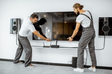 Two professional workmen in workwear installing a large TV monitor and audio system in the white...