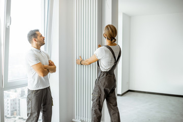 Fototapeta premium Two professional workmen in workwear installing decorative radiator in the white living room, finishing the repair in the apartment